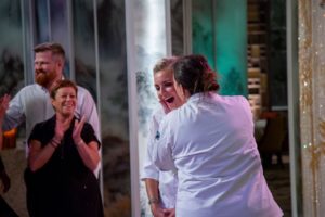 two people in white chef shirts, one with a shocked face, they other kissing her cheeck